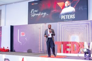 INFINION CEO Olufunsho Peters Headlines TEDxMaitama 2026 - INFINION Technologies - INFINION Technologies A man in a suit giving a TEDx talk on a stage with a large screen behind him that reads Now Speaking and features graphics and the TEDx logo on the right edge.